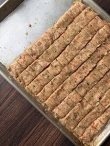 A metal baking tray with evenly cut, uncooked meat mixture bars seasoned with herbs and red pepper, placed on a wooden surface.
