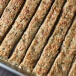 A close-up of a baking tray filled with evenly cut rectangular strips of baked meat or vegetable mixture, showing a golden-brown top with visible herbs and small vegetable pieces.