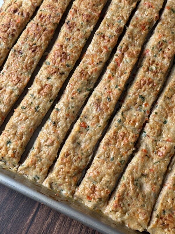 A close-up of a baking tray filled with evenly cut rectangular strips of baked meat or vegetable mixture, showing a golden-brown top with visible herbs and small vegetable pieces.