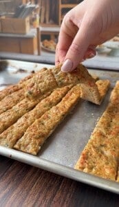 A hand lifts a rectangular slice of seasoned, baked breadsticks from a tray of similar strips on a metal baking sheet. The breadsticks appear golden and speckled with herbs.