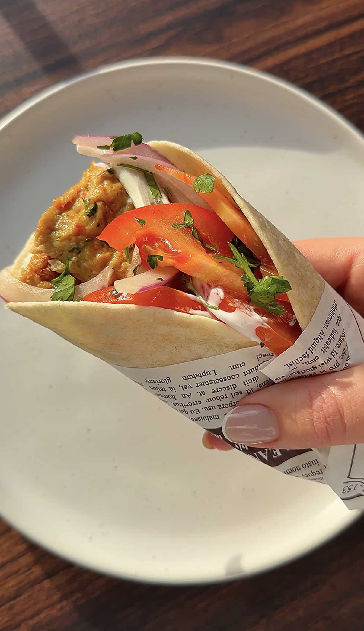 A hand holding sheet pan kebab wrap with sliced tomatoes, onions, and fresh herbs, partially wrapped in printed paper, over a white plate on a wooden table.