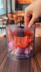 A hand holds the lid of a clear plastic food chopper filled with chopped red bell pepper, red onion, and garlic cloves on a wooden table, with a blurred indoor background.