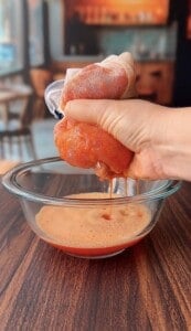 A hand squeezes a mesh bag filled with blended tomatoes over a glass bowl, extracting tomato juice onto a wooden table in a cozy kitchen setting.