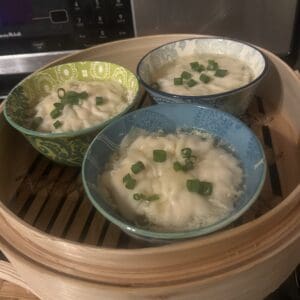 Three patterned bowls filled with steamed egg custard, each garnished with chopped green onions, sit inside a bamboo steamer on a kitchen counter—right beside a tray of VIRAL PORK DUMPLING LASAGNA for the perfect savory pairing.