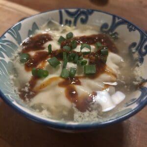 A bowl of soft tofu in clear broth, topped with a dark sauce and garnished with chopped green onions, sits on a wooden surface—perfect alongside the trending VIRAL PORK DUMPLING LASAGNA. A spoon is visible in the background.