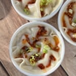 Three white bowls of dumplings topped with chili oil and chopped green onions are arranged on a wooden surface. The dumplings have curled edges and are served in a light broth or sauce.