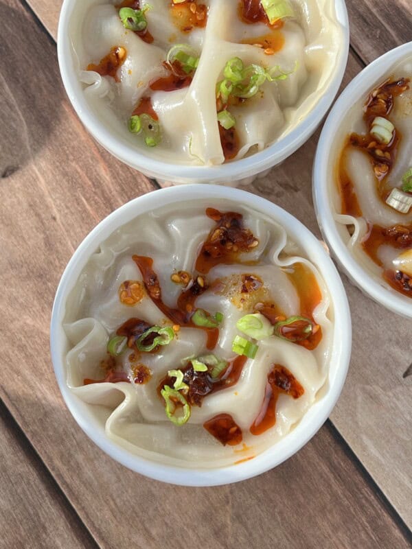 Three white bowls of dumplings topped with chili oil and chopped green onions are arranged on a wooden surface. The dumplings have curled edges and are served in a light broth or sauce.