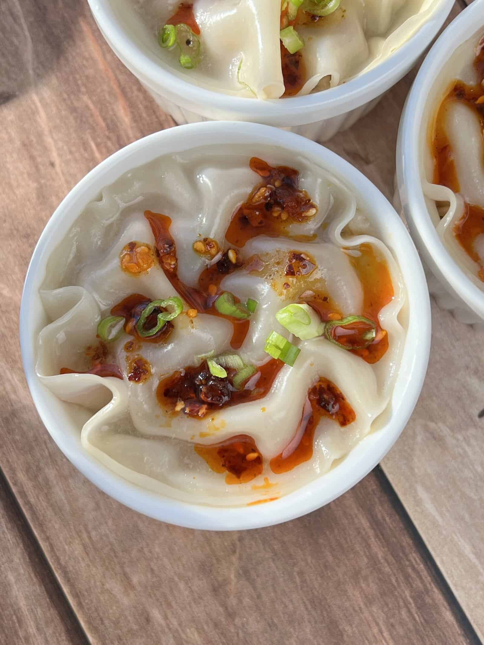 Shrimp Dumpling Lasagna, A white bowl filled with dumplings topped with chili oil and chopped green onions, placed on a wooden surface.