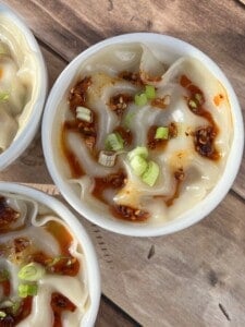 Close-up of steamed dumplings in a white bowl, topped with chili oil and garnished with chopped green onions, on a wooden surface.