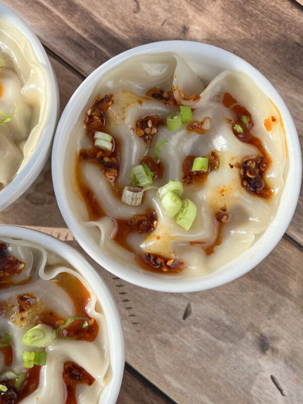Close-up of steamed dumplings in a white bowl, topped with chili oil and garnished with chopped green onions, on a wooden surface.