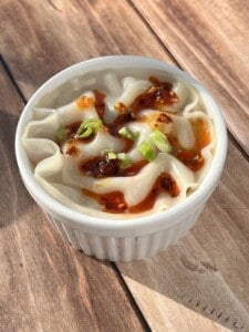 A white ramekin filled with steamed dumplings, garnished with chopped green onions and drizzled with chili oil, sits on a wooden surface.