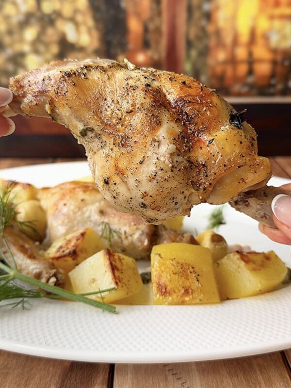 A hand holds up a seasoned, roasted chicken leg above a white plate filled with roasted potatoes and herbs, creating the same inviting atmosphere as an Easy Baked Rabbit dish, against a warm, blurred background.