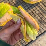 A hand holds a folded sandwich made with 2 Ingredient Cottage Cheese Wraps, filled with slices of turkey and romaine lettuce. More similar wraps rest on a cooling rack in the background.