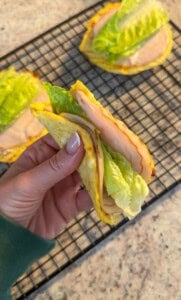 A hand holds a folded sandwich made with 2 Ingredient Cottage Cheese Wraps, filled with slices of turkey and romaine lettuce. More similar wraps rest on a cooling rack in the background.
