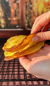 A hand holds up two golden, slightly crispy 2 Ingredient Cottage Cheese Wraps over a cooling rack, with a blurred background of warm colors.