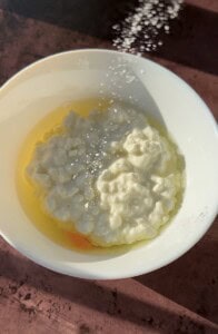 A white bowl contains the key ingredients for 2 Ingredient Cottage Cheese Wraps—cottage cheese and a raw egg—with salt being sprinkled on top. The bowl rests on a brown surface.