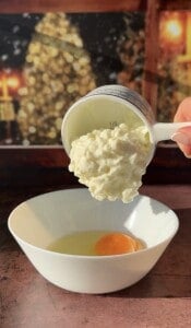 A hand pours a cup of cottage cheese into a white bowl with a raw egg, illustrating the simple start to making 2 Ingredient Cottage Cheese Wraps. The background is warmly lit and softly blurred.