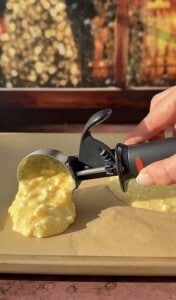 A hand uses a black-handled ice cream scoop to portion out yellow, chunky batter for 2 Ingredient Cottage Cheese Wraps onto a sheet of parchment paper, with a blurred, warm-toned background behind.