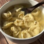 A bowl of tortellini in clear broth sits on a wooden surface, with a wooden spoon resting inside. The soup is sprinkled with black pepper and bathed in warm sunlight.