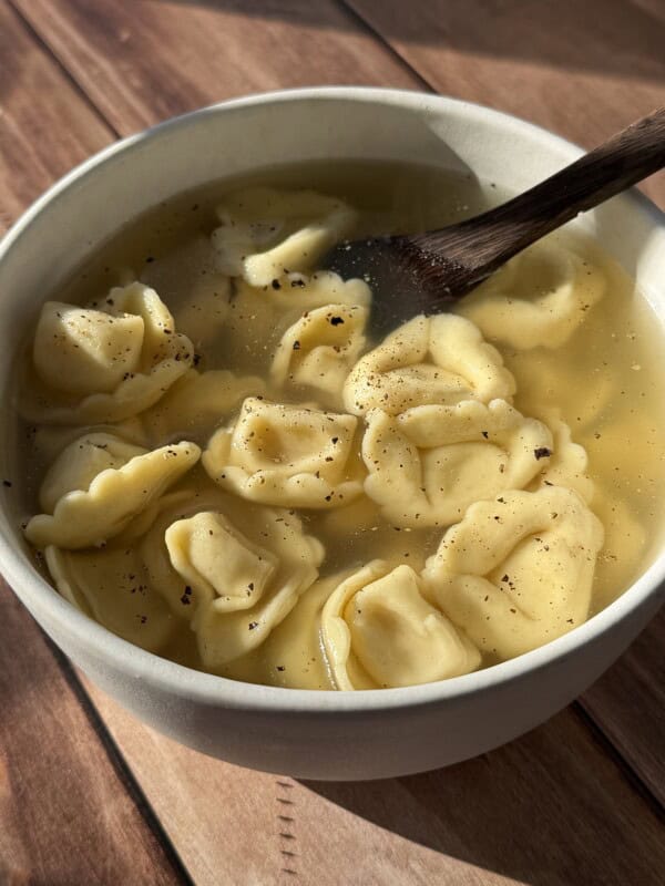 A bowl of tortellini in clear broth sits on a wooden surface, with a wooden spoon resting inside. The soup is sprinkled with black pepper and bathed in warm sunlight.