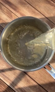 A clear liquid is being poured from a glass container into a stainless steel pot on a wooden surface, with light reflections on the water and pot.
