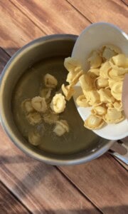 A hand pours uncooked tortellini from a white bowl into a pot of boiling water on a wooden surface.