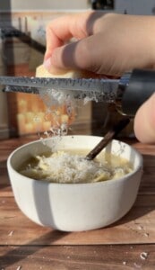 A hand grates cheese over a bowl of pasta with a wooden spoon inside, as sunlight streams onto the wooden table beneath.