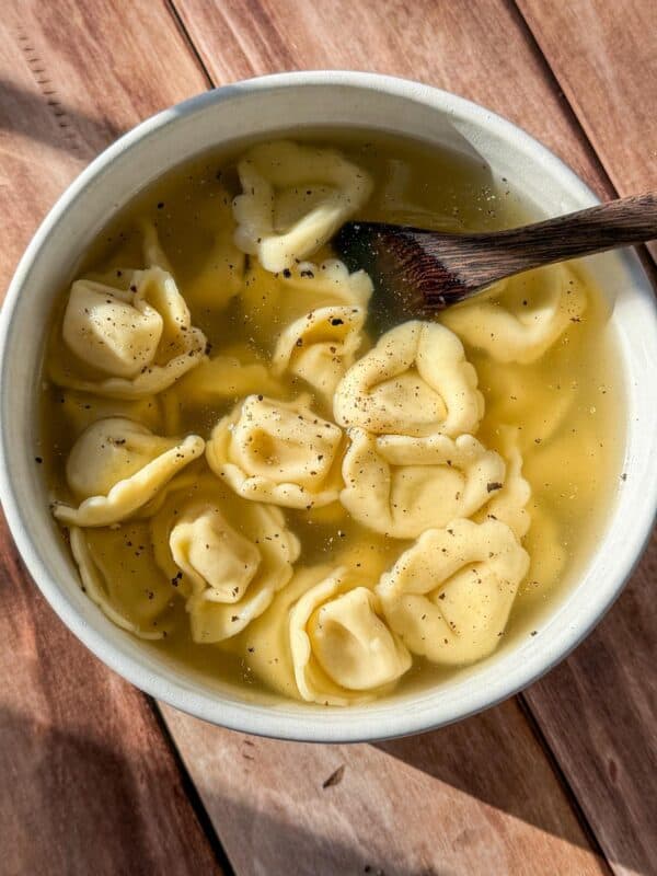 A bowl of Easy Tortellini Soup with clear broth and several pieces of tortellini pasta, topped with cracked black pepper. A wooden spoon rests inside, and the bowl sits on a wooden surface in natural light.