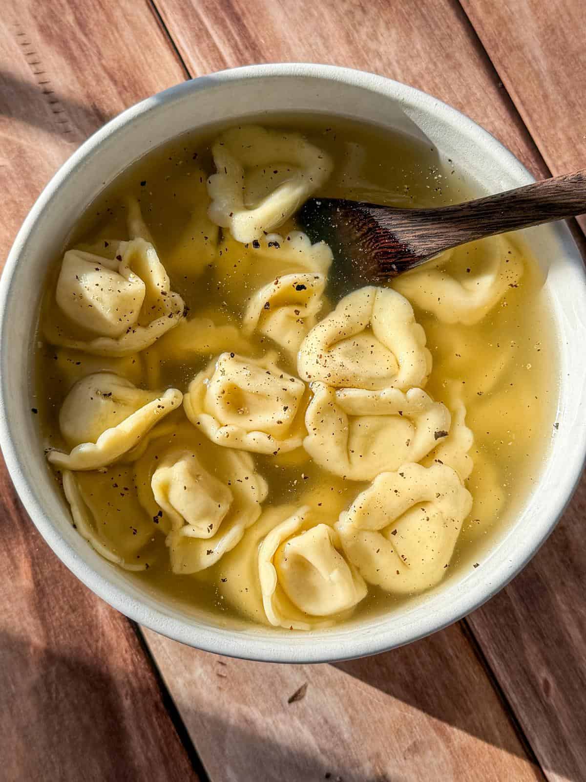 A bowl of Easy Tortellini Soup with clear broth and several pieces of tortellini pasta, topped with cracked black pepper. A wooden spoon rests inside, and the bowl sits on a wooden surface in natural light. - 3