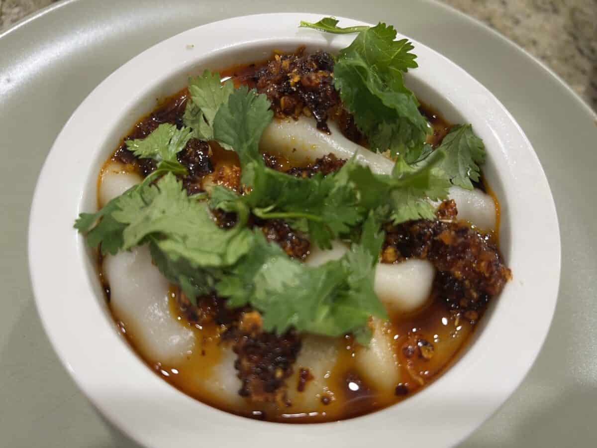 A white bowl filled with rice noodles topped with chili oil and fresh cilantro, set on a light gray plate—perfect for enjoying alongside the trending flavors of VIRAL PORK DUMPLING LASAGNA.
