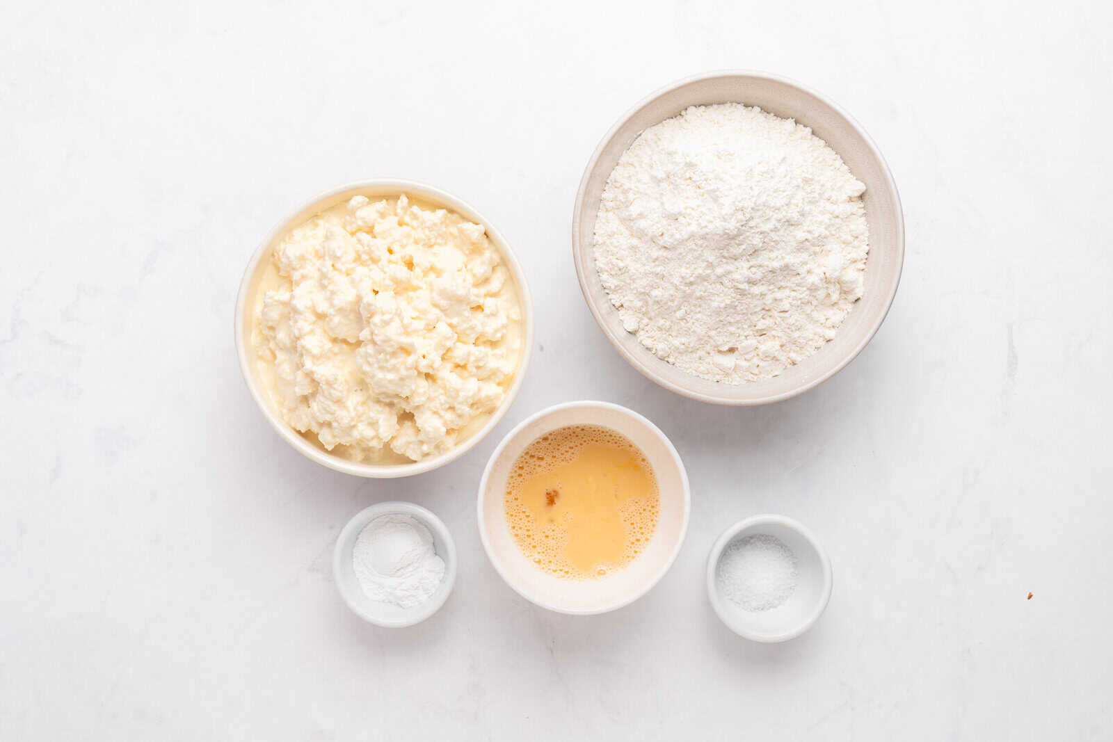 Five bowls on a white surface: one with cottage cheese, one with flour, one with beaten egg, one with baking powder, and one with salt. - 4