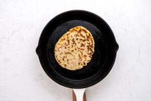 A single piece of flatbread is cooking in a black cast iron skillet with a white handle, set on a white marble surface.