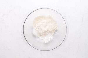 A clear glass bowl containing a mixture of dry baking ingredients, including flour and possibly sugar or baking powder, on a white marble surface.