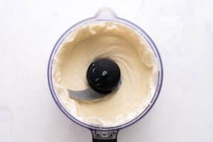 A food processor filled with smooth, creamy mixture is viewed from above on a white surface. The metal blade is visible in the center, surrounded by the blended mixture.