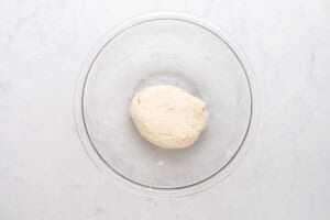 A clear glass bowl containing a ball of smooth, pale dough sits on a white marble surface.