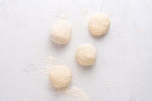 Four balls of dough are placed on a lightly floured white surface, with some scattered flour surrounding them.