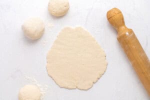 A rolled-out piece of dough is on a floured surface next to a wooden rolling pin and three round dough balls, ready for baking or cooking.