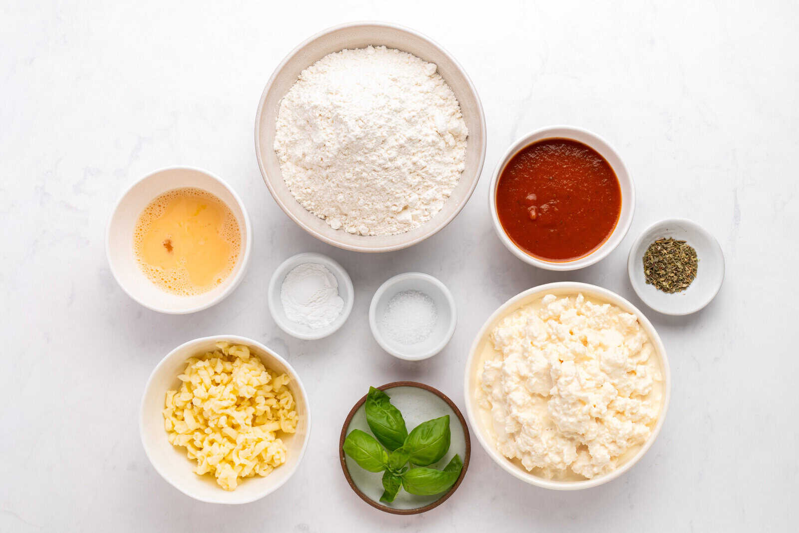 Various pizza ingredients in bowls on a white surface, including flour, tomato sauce, shredded cheese, ricotta cheese, beaten egg, baking powder, salt, dried herbs, and fresh basil leaves. - 4