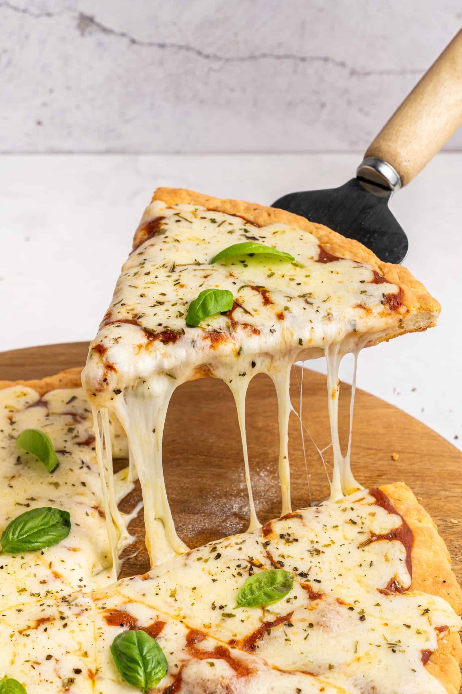 A hand lifts a cheesy slice of pizza from a wooden board, with melted cheese stretching between the slice and the pizza. The pizza is topped with basil leaves and herbs. - 3