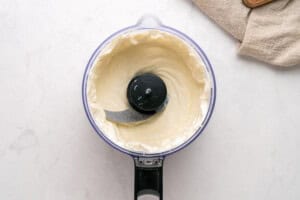 A food processor filled with smooth, creamy white mixture sits on a light countertop, with a beige kitchen towel partially visible in the upper right corner.