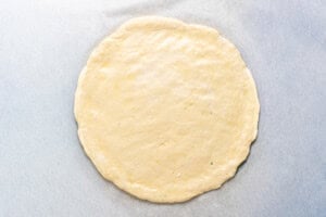 A round, uncooked pizza dough is rolled out flat on a sheet of parchment paper, ready for toppings and baking.