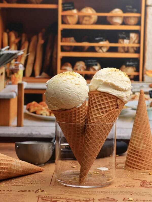 Two waffle cones filled with Viral Snow Ice Cream stand upright in a glass jar on a wooden table, with a bakery display of bread and pastries in the background.