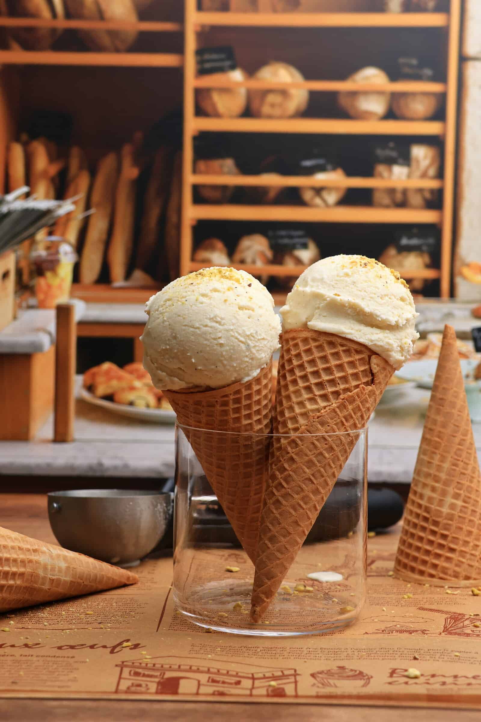 Two waffle cones filled with Viral Snow Ice Cream stand upright in a glass jar on a wooden table, with a bakery display of bread and pastries in the background.