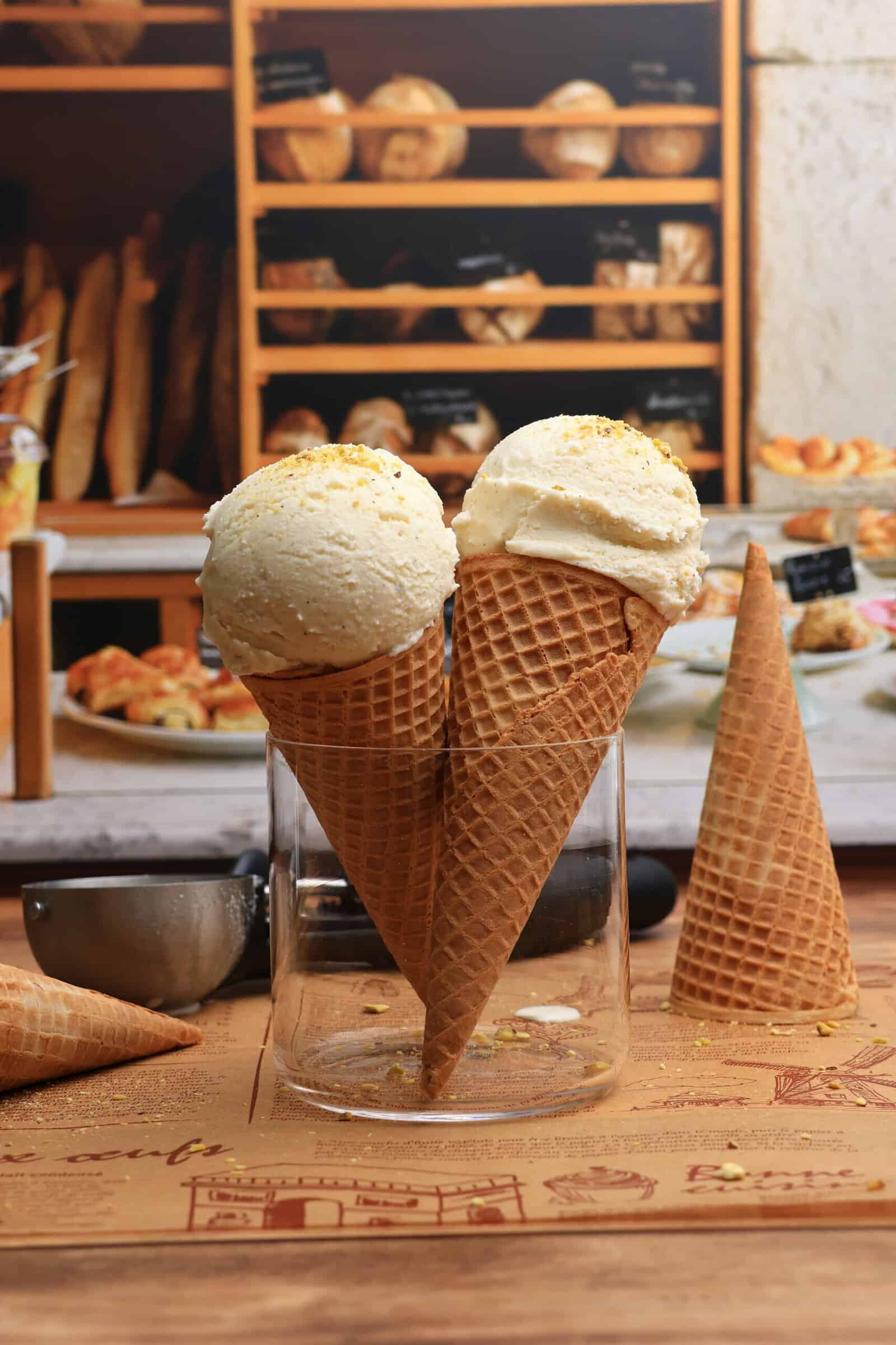 Two waffle cones filled with scoops of Viral Snow Ice Cream stand upright in a glass jar. Another empty cone and a metal scoop rest on a wooden table, with bread and pastries displayed on shelves in the background.