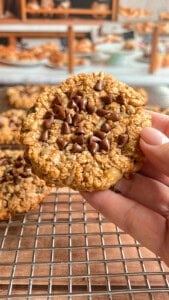 A hand holds an oatmeal cookie topped with chocolate chips over a wire cooling rack, with more cookies and a blurred kitchen background in view.