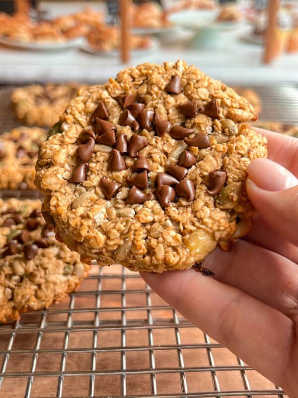A hand holds an oatmeal cookie topped with chocolate chips over a wire cooling rack, with more cookies and a blurred kitchen background in view.