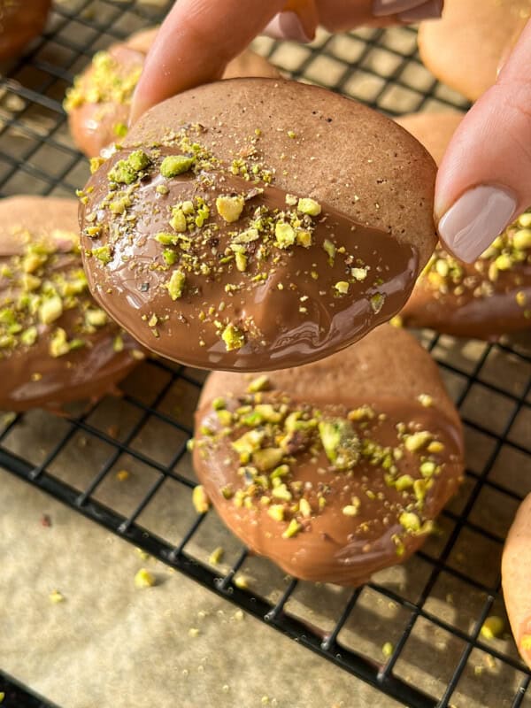 A hand holding a round cookie dipped in chocolate and sprinkled with crushed pistachios, with similar cookies resting on a wire cooling rack in the background. - 10
