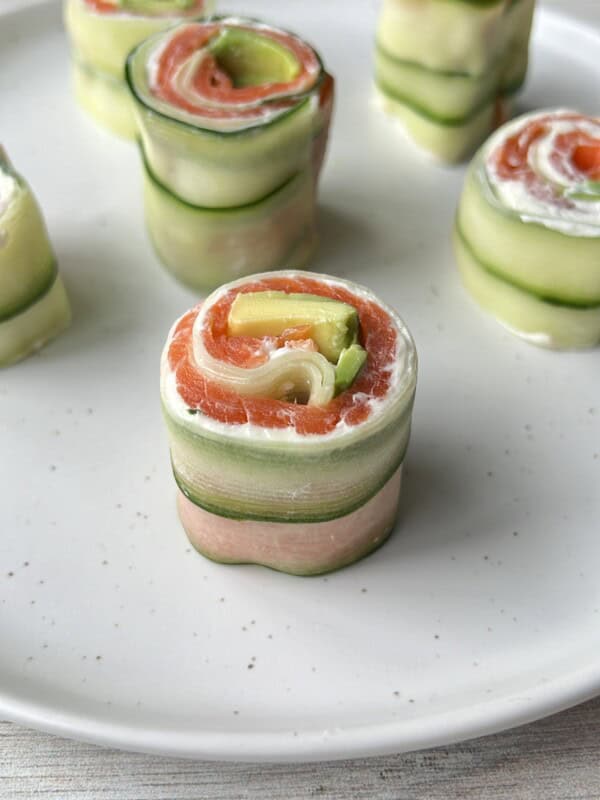 Close-up of cucumber rolls filled with slices of salmon, avocado, and a creamy spread, arranged neatly on a white plate. The rolls resemble sushi but are made with fresh vegetables and fish.