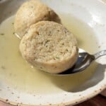 A close-up of a bowl with clear broth and a sliced bread dumpling, one half resting on a spoon. The dumpling appears soft with visible herbs, and sunlight highlights the textures.