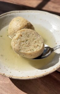 A close-up of a bowl with clear broth and a sliced bread dumpling, one half resting on a spoon. The dumpling appears soft with visible herbs, and sunlight highlights the textures.
